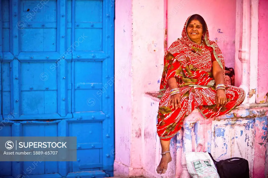 A Woman In A Red Sari Sits Beside A Blue Door, Haridwar India
