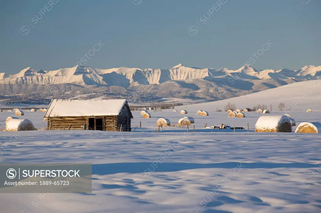 snow covered log cabin and hay bales in a field with snow covered mountains, okotoks, alberta, canada