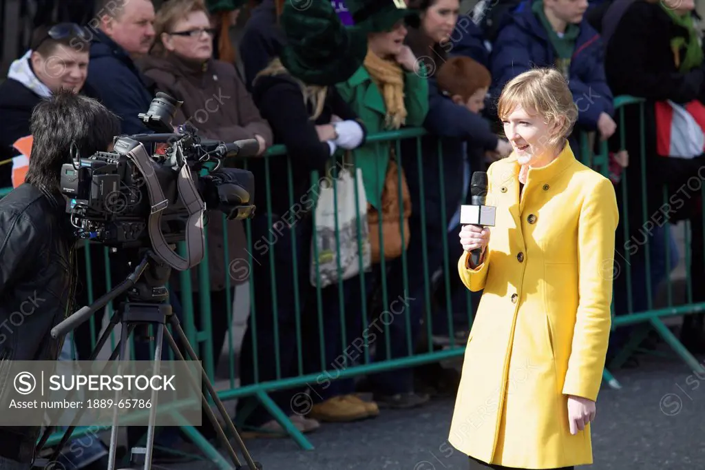 a woman news reporter reporting in front of a camera beside the crowd waiting at the saint patrick´s day parade, dublin ireland
