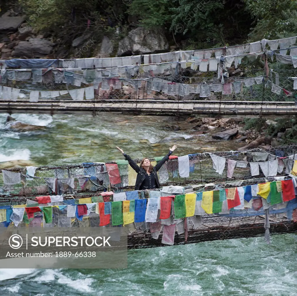 a woman with raised arms on an iron suspension bridge lined with prayer flags, tamchhog lhakhang bhutan