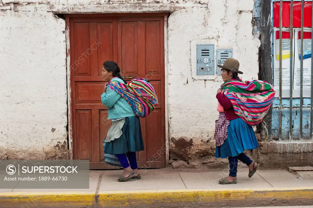 Two Women Walking On A Sidewalk Carrying Blanket Bundles From The Market, Cusco Peru