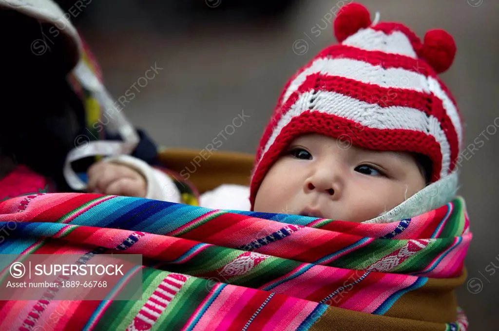 A Baby Wearing A Red And White Striped Hat Being Carried In A Colorful Bundle, Cusco Peru