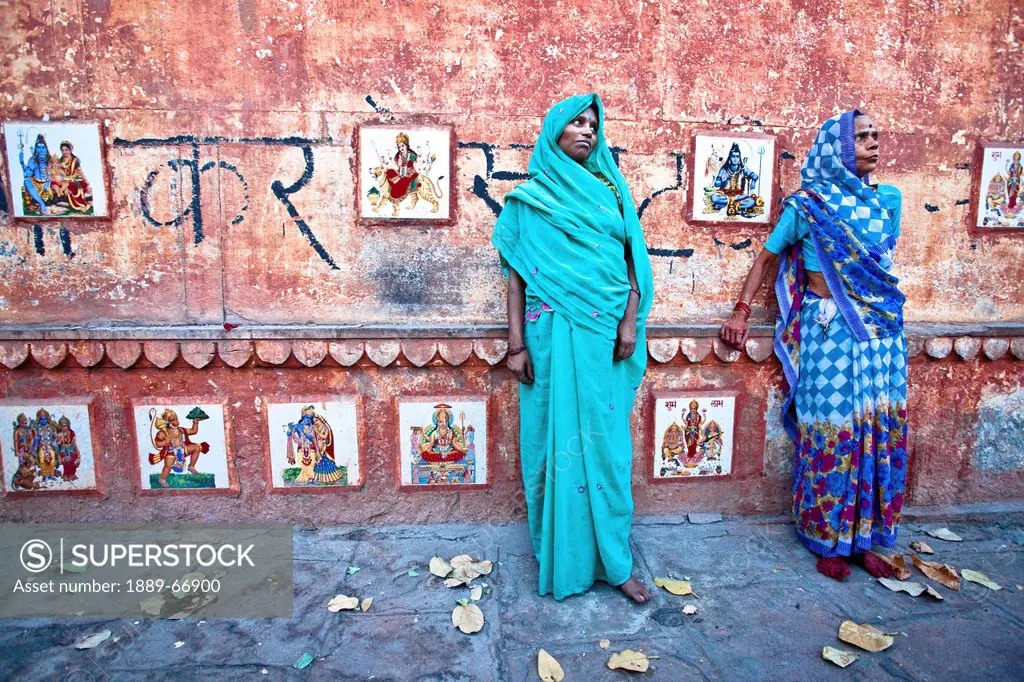 Two Women In Saris Standing In The Street Against A Wall Lined With Pictures Of Hindu Gods, Varanasi India