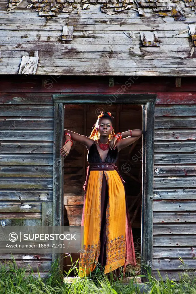 a young woman dressed in colourful clothing and make_up stands confidently in the doorway of a weathered shed, leduc, alberta, canada