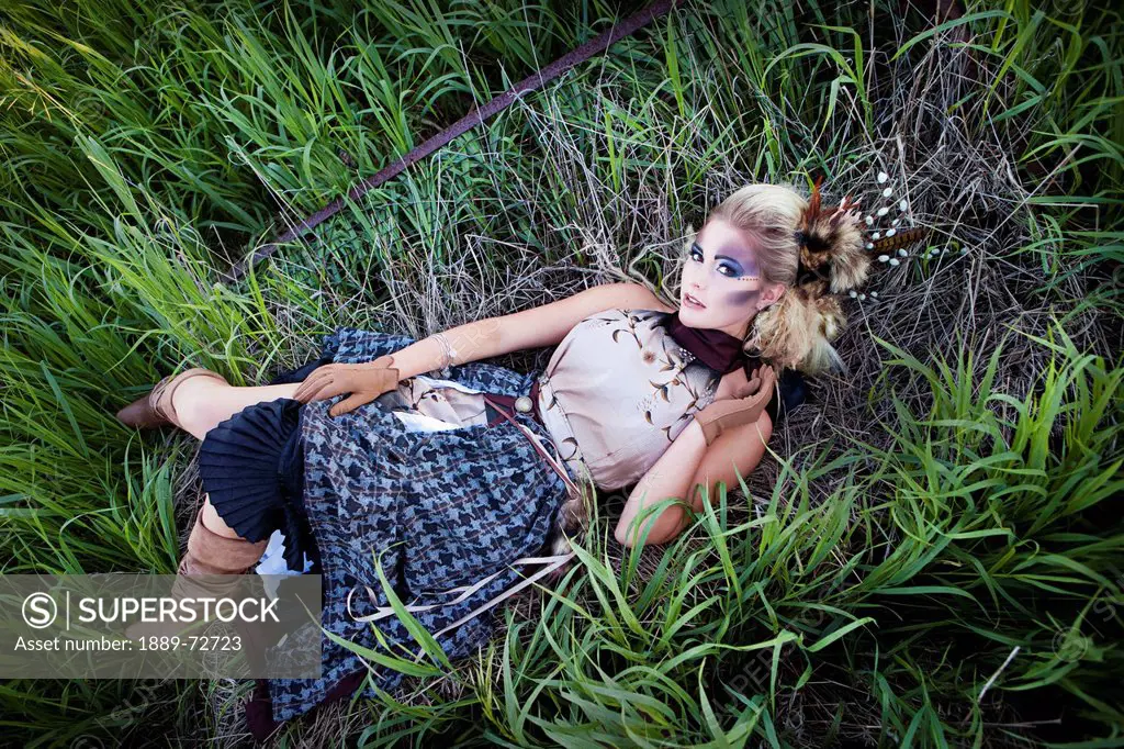 a young woman with pronounced makeup and a feather hair accessory laying in the grass, nisku, alberta, canada