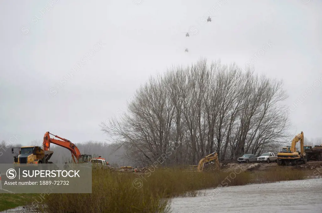 tractors and trucks working on the riverbank to prevent flooding with army helicopters approaching in the air, newton manitoba canada
