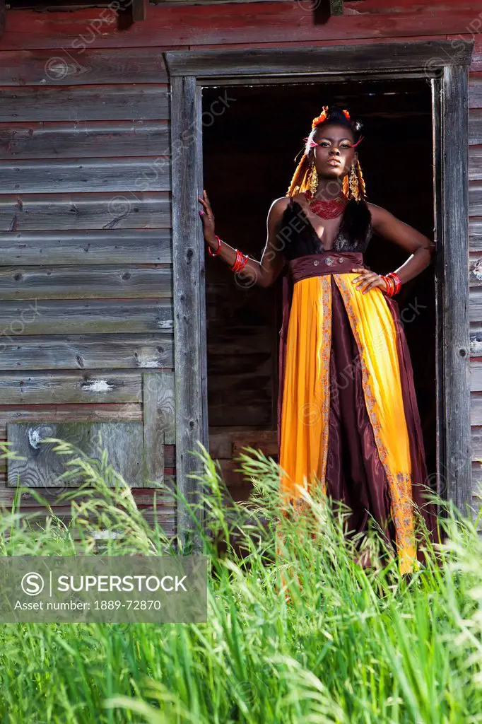 a young woman in a colourful dress, makeup and accessories standing in the doorway of a weathered shed, leduc, alberta, canada