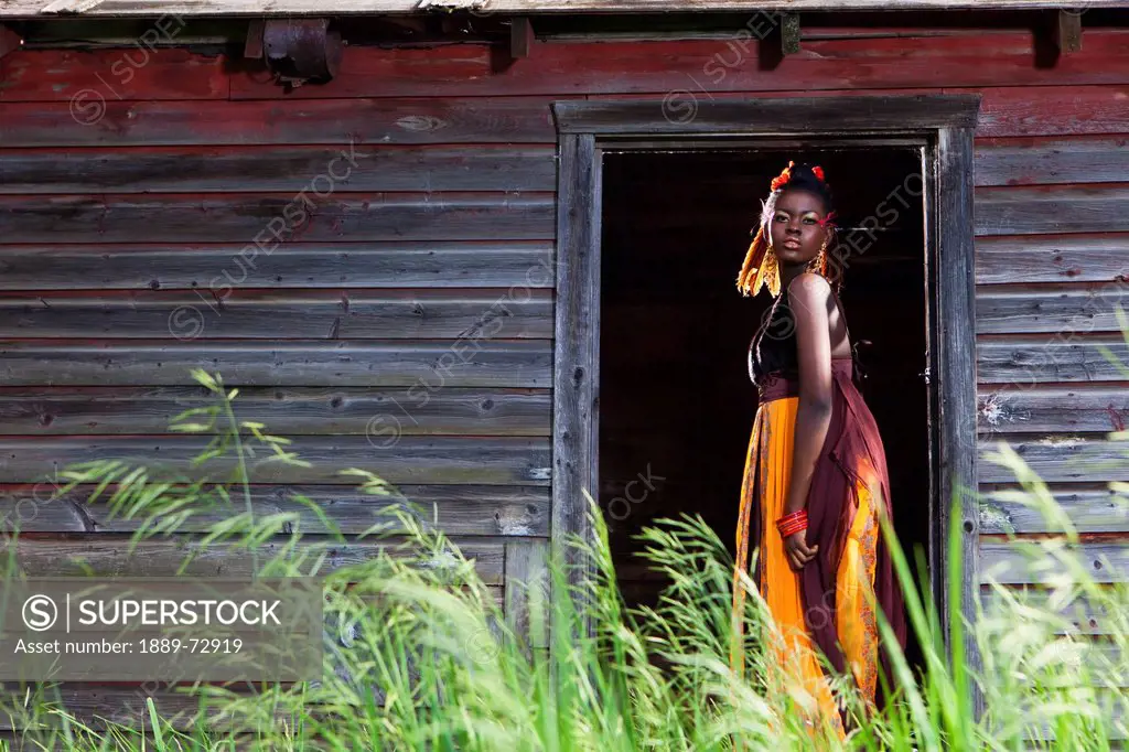 a young woman in a colourful dress, makeup and accessories standing in the doorway of a weathered shed, leduc, alberta, canada