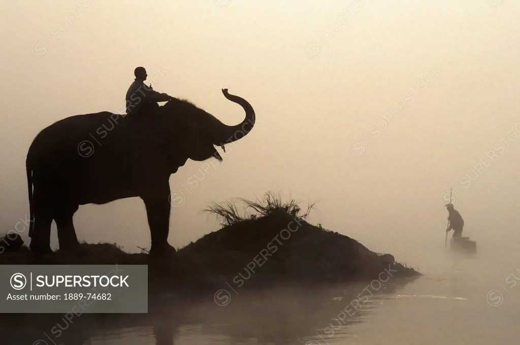 An Elephant With Its Mahout Stand At The Edge Of The Rapti River Near Sauraha And Chitwan National Park As A Man Pushes His Dugout Canoe Along The Riv...