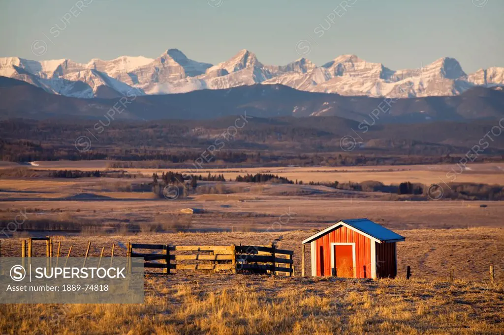Warmly Lit Red Shack In Field With Wooden Fence And Mountains In The Background With Blue Sky At Sunrise South Of Cochrane, Alberta Canada