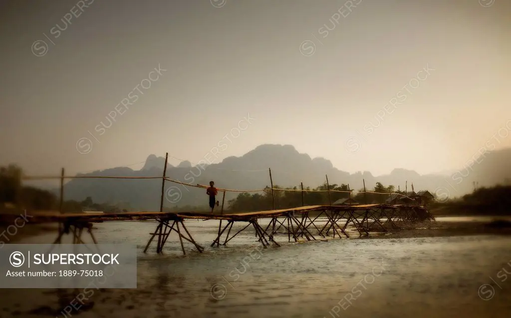 Boy running across a wooden bridge on a river with mountains in the background, vang vieng laos