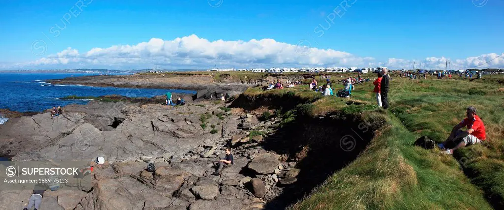 People sitting on the rocks and grass along the coast at hook lighthouse looking out at the ocean, county wexford ireland