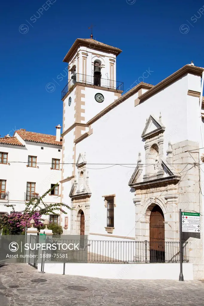 White church building with a clock and cross on a tower against a blue sky, grazalema andalusia spain