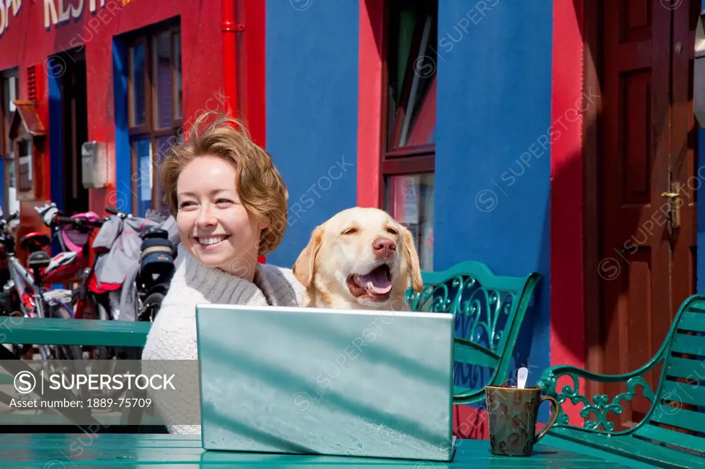 A woman sits at a table with her dog and a laptop computer, allihies county cork ireland