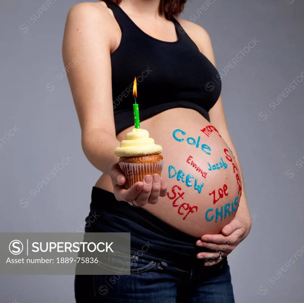 A pregnant woman with baby names written all over her bare belly holding a cupcake with lit candle, edmonton alberta canada