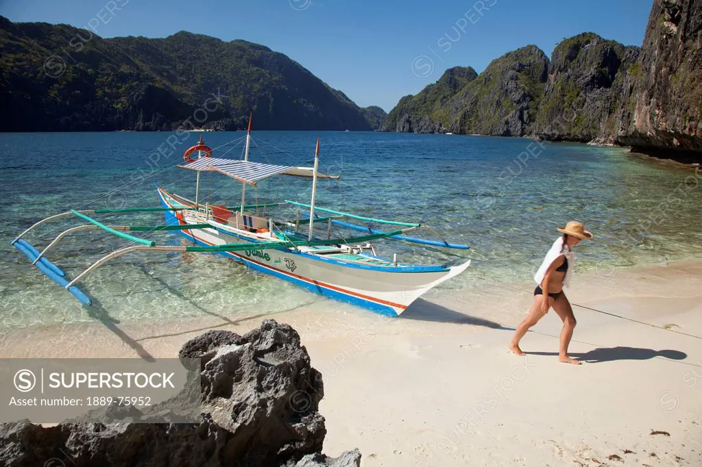 A Woman Tourist Wearing A Sun Hat And Bikini Walks Past A Bangka Boat Of Matinloc Island Near El Nido And Corong Corong, Bacuit Archipelago Palawan Ph...