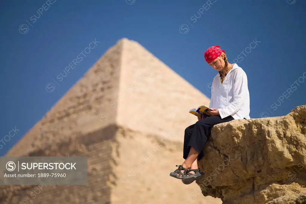 A Female Tourist Reads A Guide Book While Sitting In Front Of The Pyramids Of Giza Near Cairo, Giza Egypt