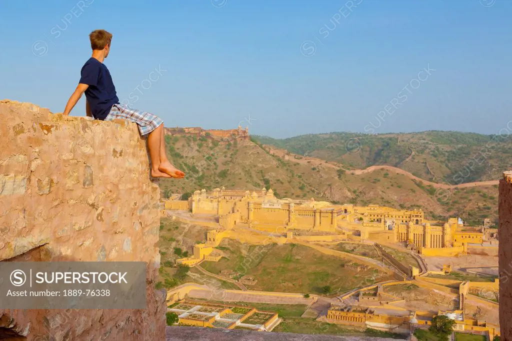 A young man sits barefoot on the top of a wall looking out over the amer fort, jaipur rajasthan india