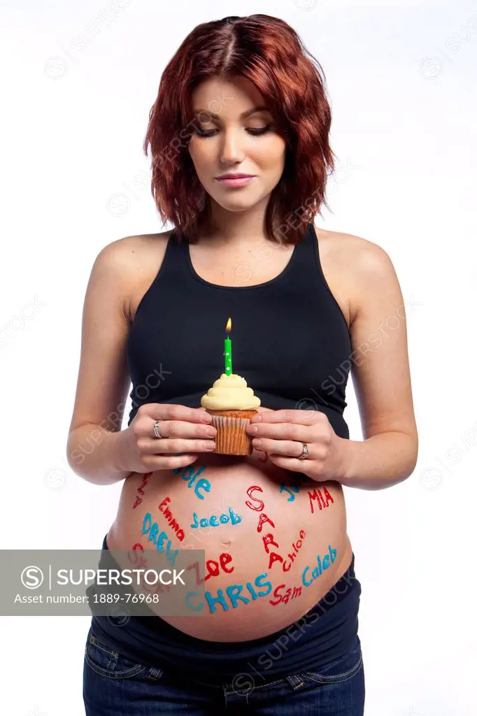 A pregnant woman with names written all over her bare belly holding a cupcake with a lit candle, edmonton alberta canada