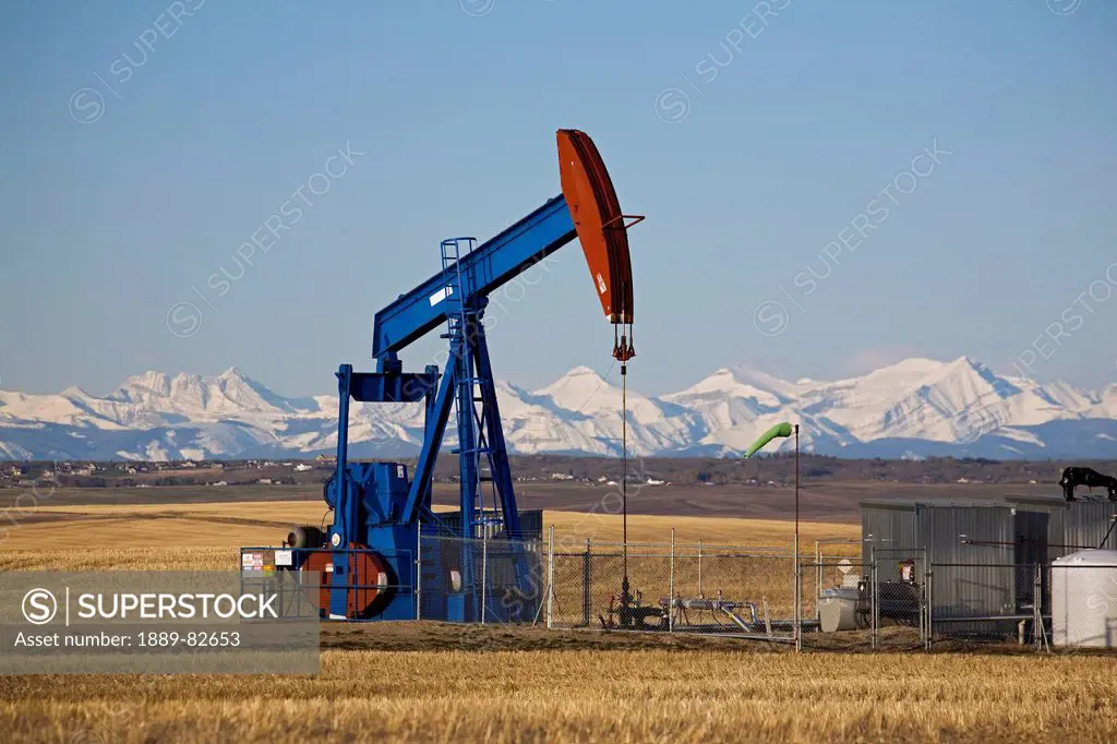 Pumpjack in a field with snow covered mountains and blue sky in the background, alberta, canada