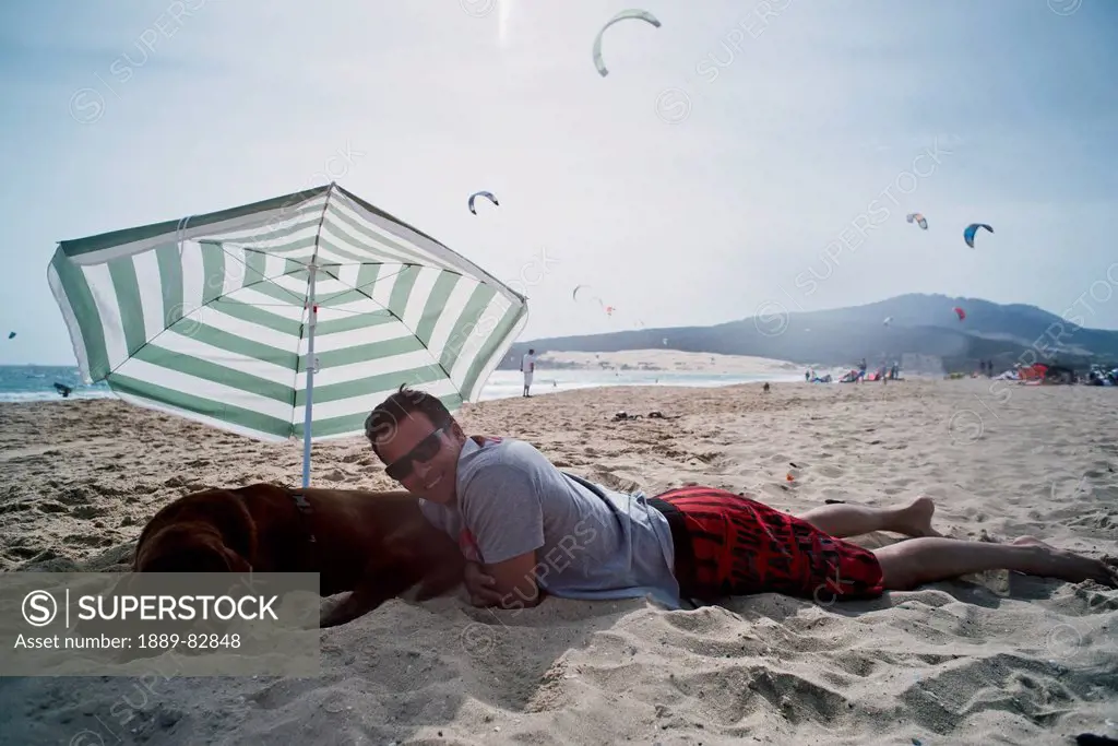 A man laying on punta paloma beach with his dog under an umbrella, tarifa cadiz andalusia spain