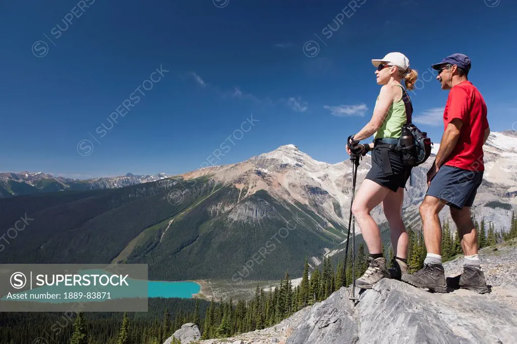 Female And Male Hikers On Top Of Rock Cliff Overlooking Lake Below, Field British Columbia Canada