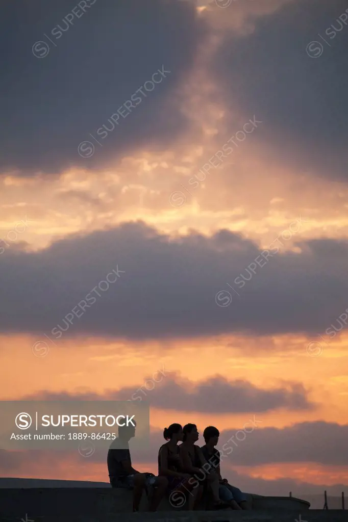 Cyprus, Four People Sitting On Ledge At Sunset; Paphos