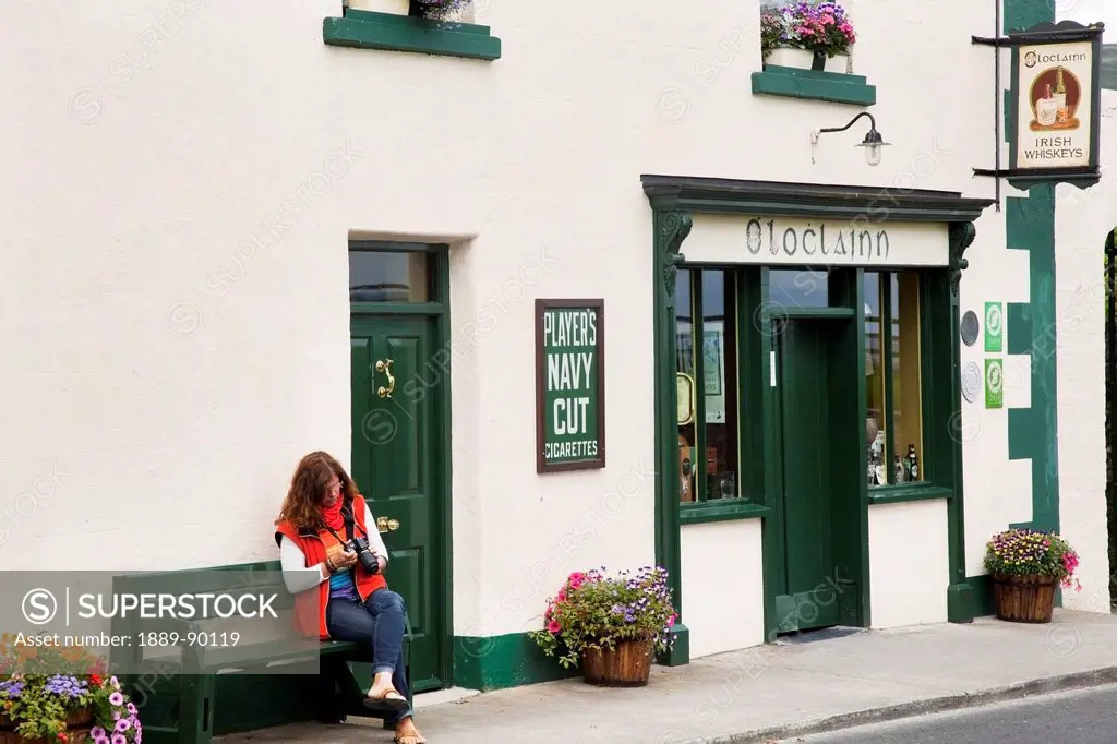 A woman sits on a bench outside a shop looking at her camera;Ballyvaughan county clare ireland