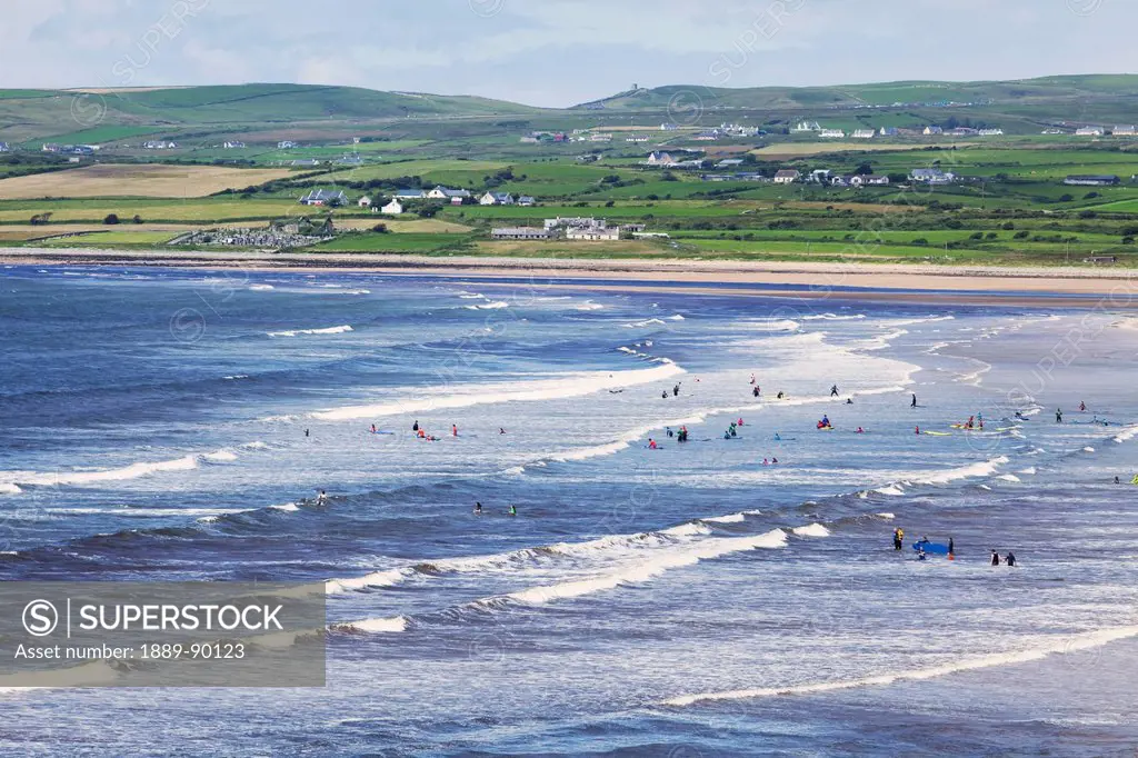 A busy beach with swimmers and surfers and houses scattered along the coast;Lahinch county clare ireland