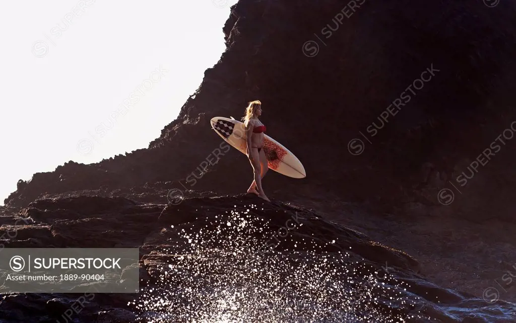 Portugal, Algarve, Carrapateira, Distant view of female surfer with surfboard standing on rock; Playa do Amado