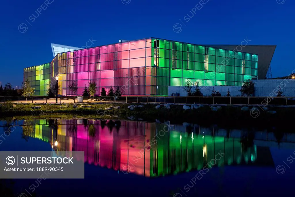 Night shot of building lit by colourful lights and reflecting in pond; Calgary, Alberta, Canada