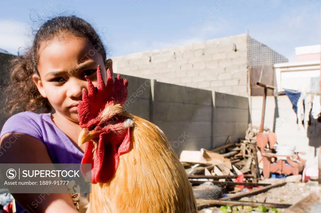 A young girl with a chicken in a yard full of debris; Mfuleni, South Africa