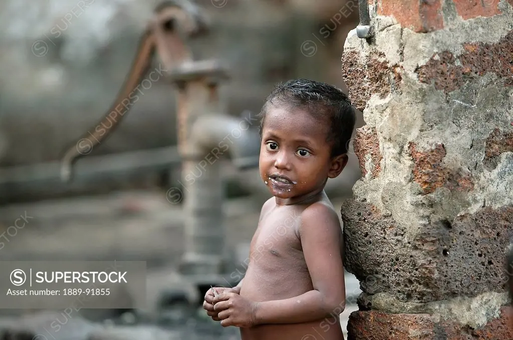 A naked child standing against a wall with a water hand pump in the background; Gopinathpur Village, India