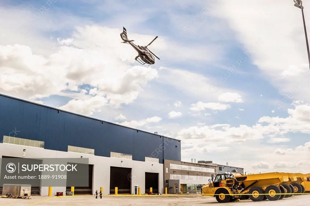 A helicopter flies above a workshop with CAT trucks parked outside; Fort McMurray, Alberta, Canada