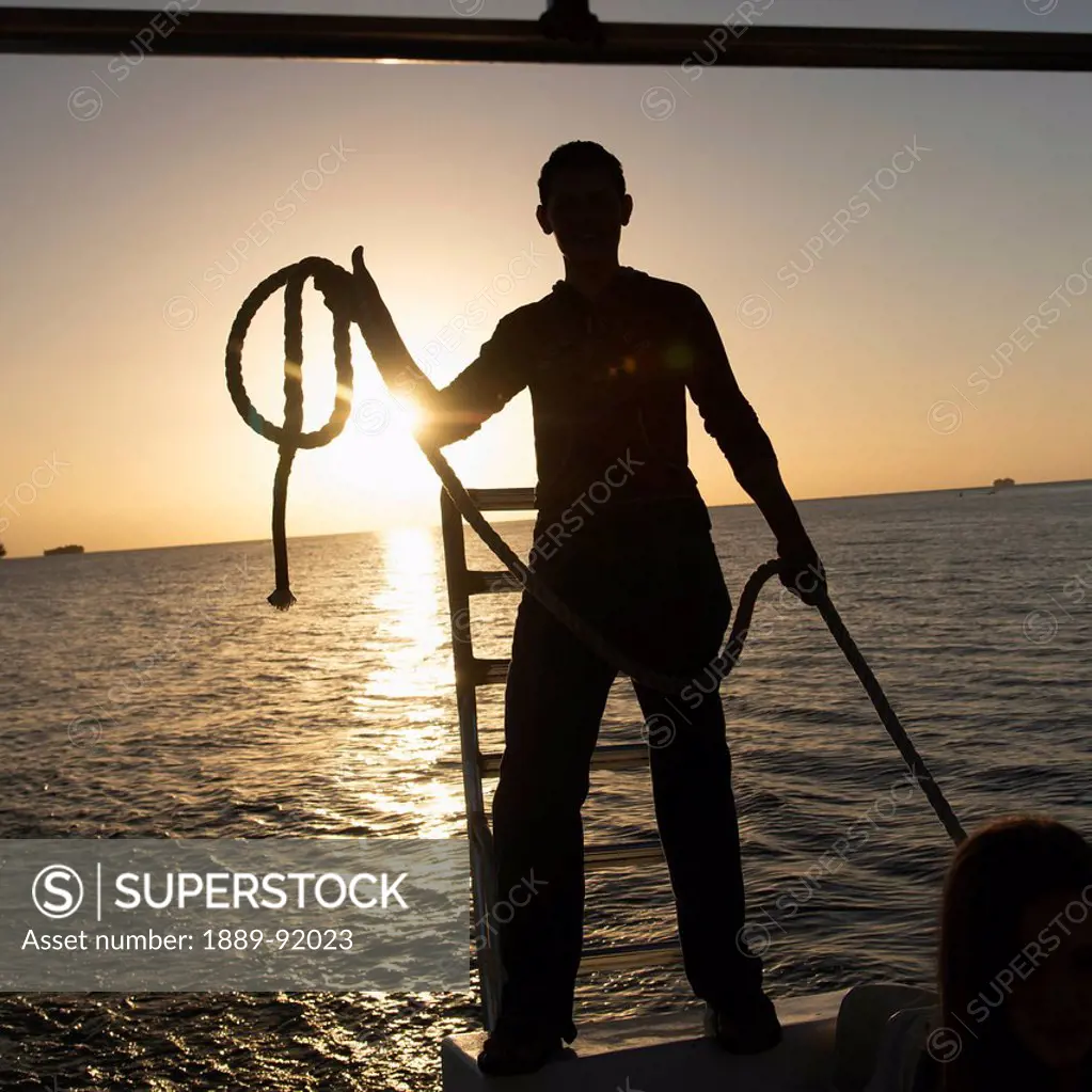 Silhouette Of A Man Standing With A Rope On The Edge Of A Boat At Sunset; Bay Islands, Honduras