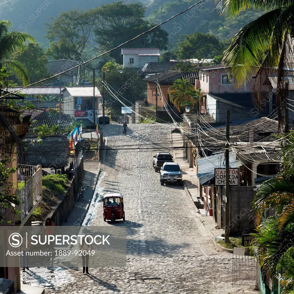 A Street With Pedestrians, Cars, Buildings And Lush Vegetation; Copan, Honduras