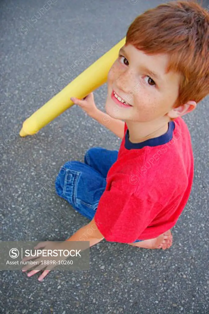 A close-up of a child sitting on asphalt with an oversized yellow crayon.