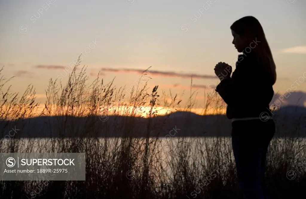 A teenage girl prays at sunset by the ocean.
