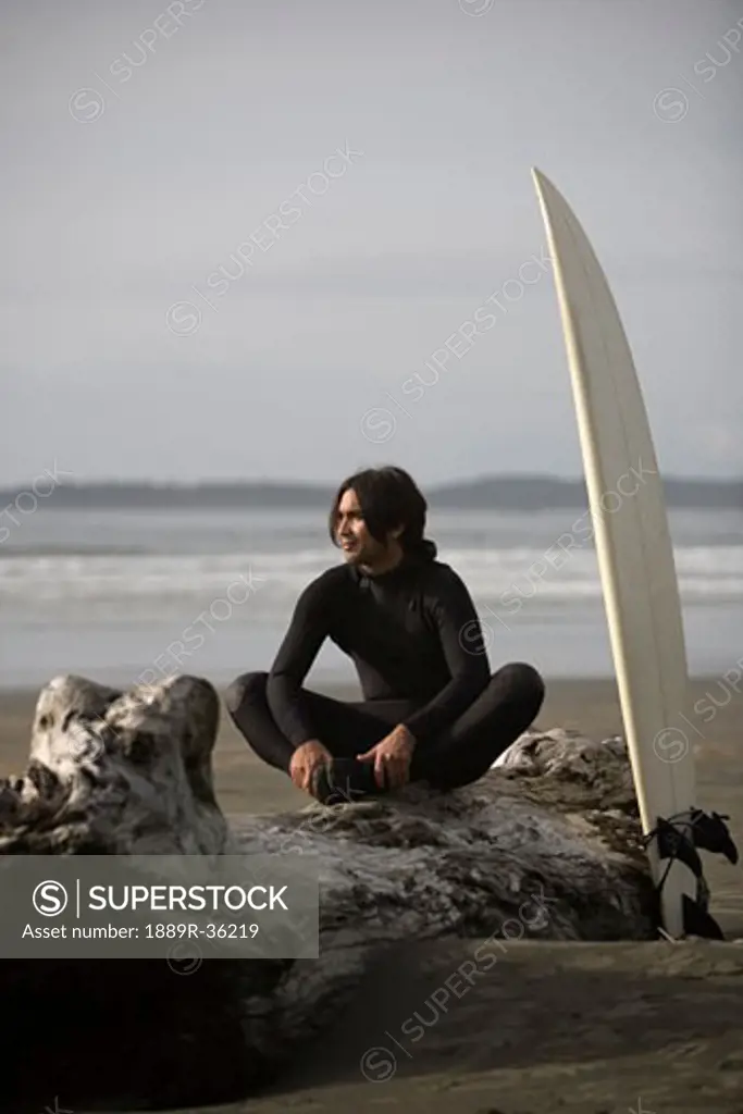 Surfer sitting on log on beach, Cox Bay near Tofino, British Columbia, Canada