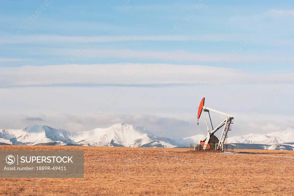 alberta, canada, pump jack in a brown field with snow covered mountains in the background