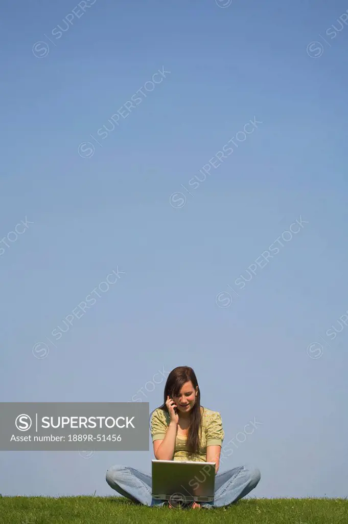 a girl working on a laptop and talking on a cellphone while sitting on the grass