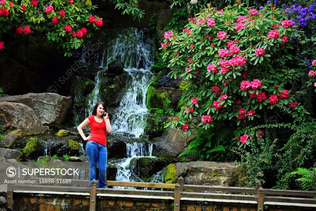 a woman talking on her cellphone while standing on a bridge with a waterfall and flowers behind her