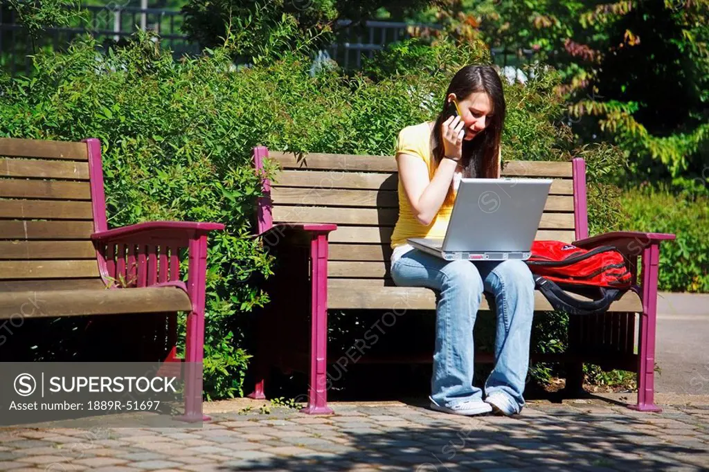 a young woman working on her laptop computer and talking on her cellphone while sitting outdoors on a park bench