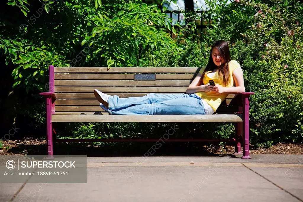 a teenage girl using her cellphone sitting on a park bench