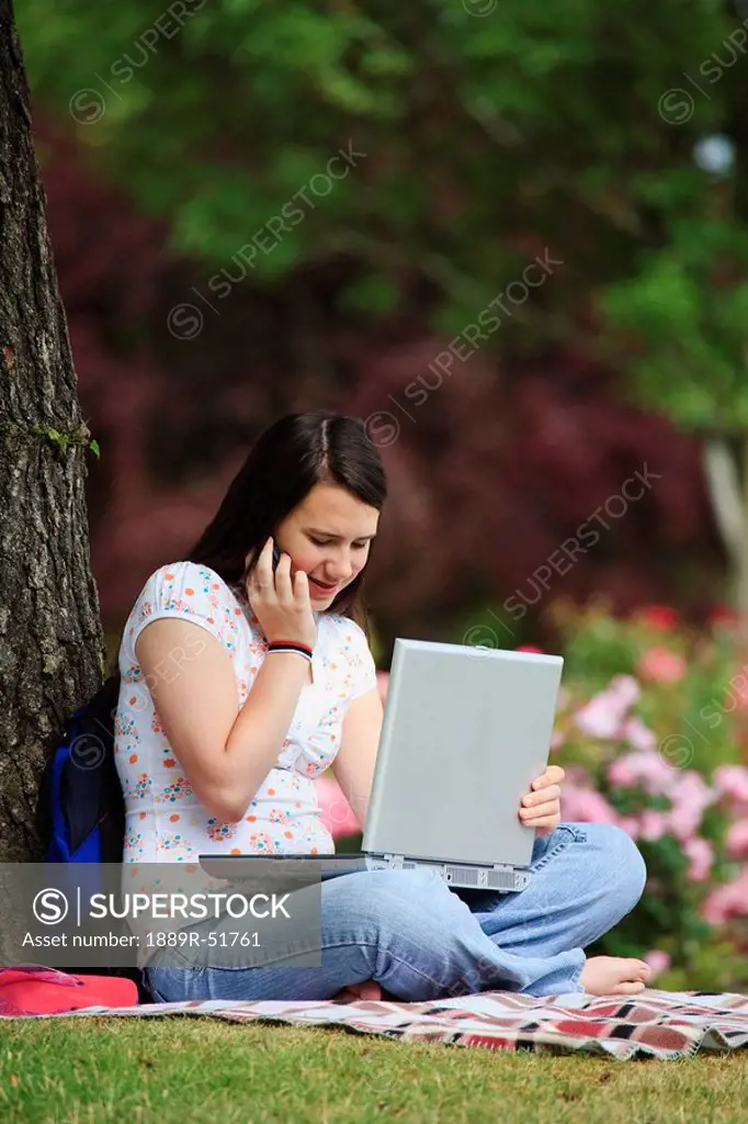 a teenage girl working on her laptop computer and talking on her cellphone under a tree