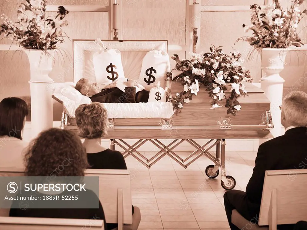 the deceased laying in a coffin at his funeral with bags of money surrounding him