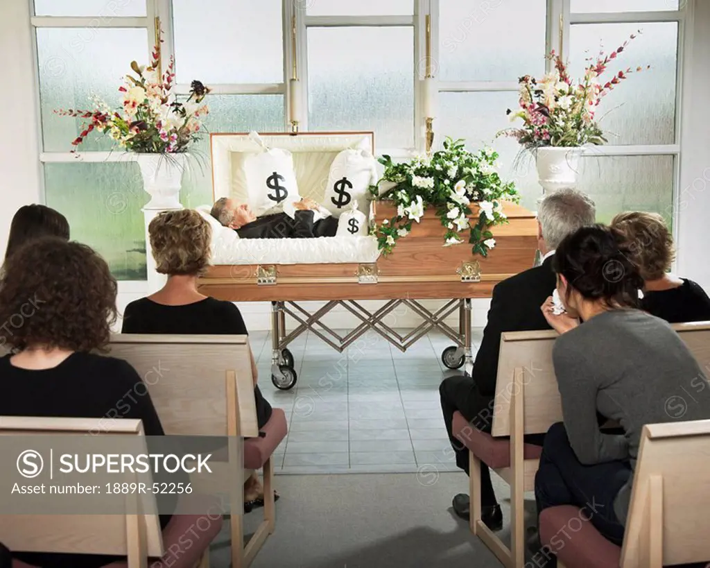 the deceased laying in a coffin at his funeral with bags of money surrounding him