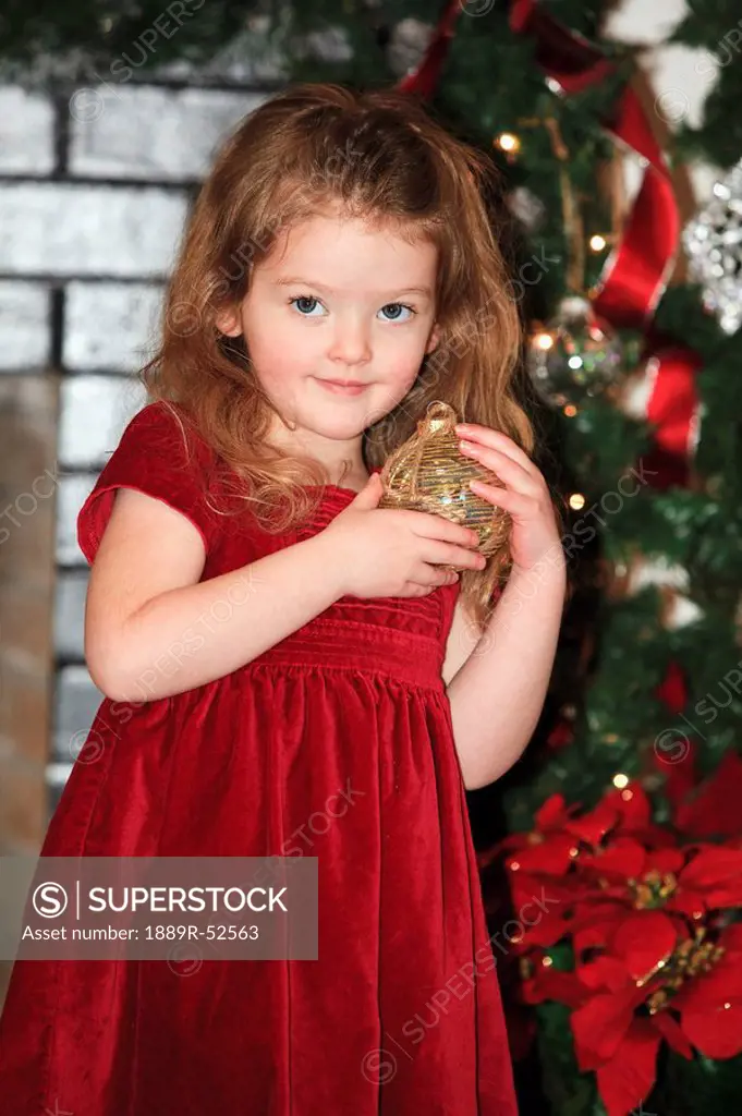 a young girl holds an ornament as she stands by the christmas tree