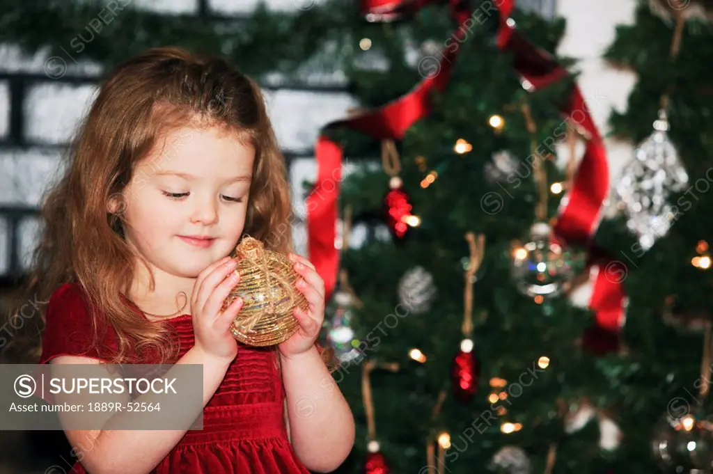 a young girl holds an ornament as she stands by the christmas tree