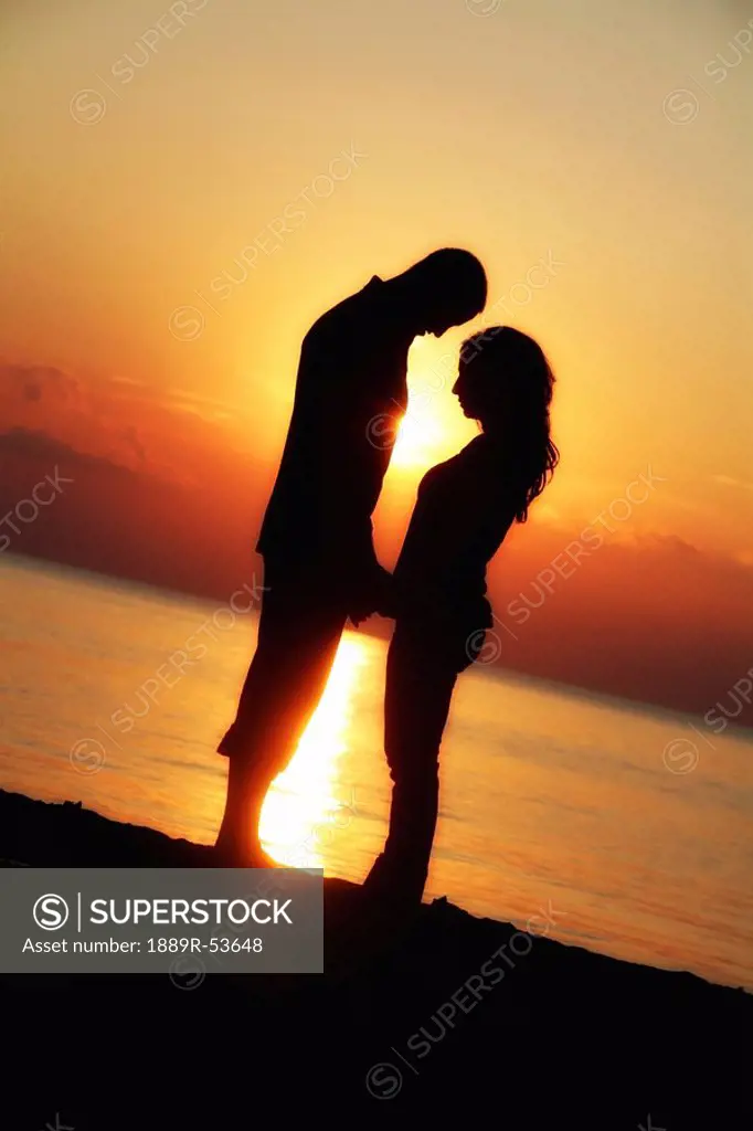 a man and woman facing each other and holding hands while standing on the beach in the sunset